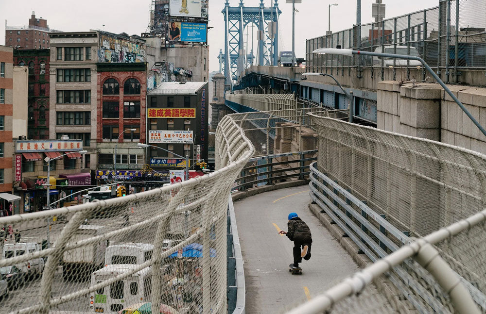 Person wearing Triple 8 protection, skateboarding on an elevated walkway with NYC cityscape in the background.